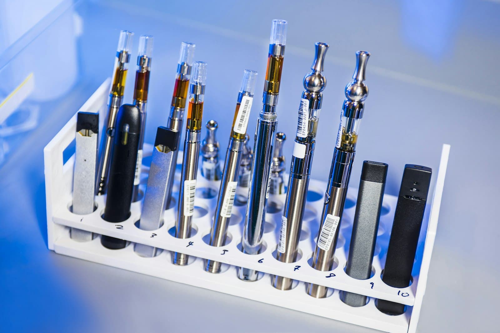 A row of different vape pens, pod devices and e-liquid cartridges lined up in a testing rack